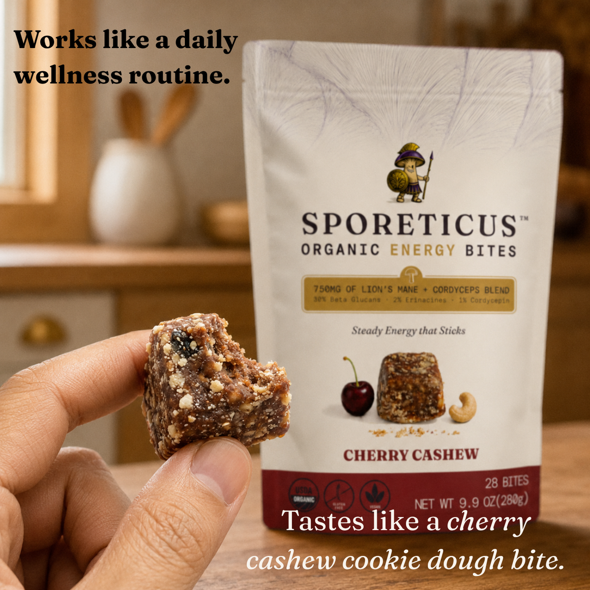 A hand holds a cherry cashew energy bite in front of a white bag of Sporeticus Organic Energy Bites. The packaging features a cartoon character and the product name 'Cherry Cashew' on a warm, blurred kitchen background.