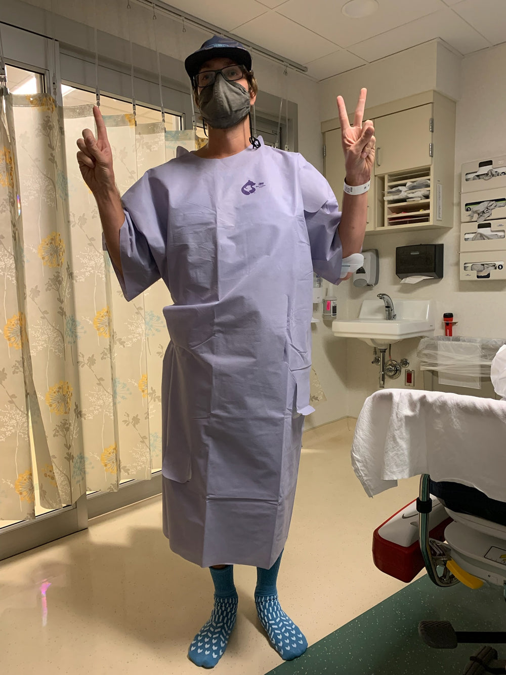 A person standing in a medical exam room wearing a light lavender surgical scrub gown, blue patterned socks, a face mask, and a cap, making peace signs with both hands.