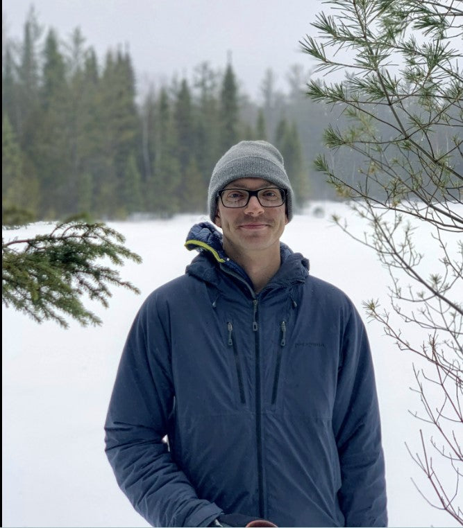 A man wearing a gray knit beanie, black eyeglasses, and a navy blue zip-up fleece jacket stands outdoors in a snowy landscape with evergreen trees in the background.