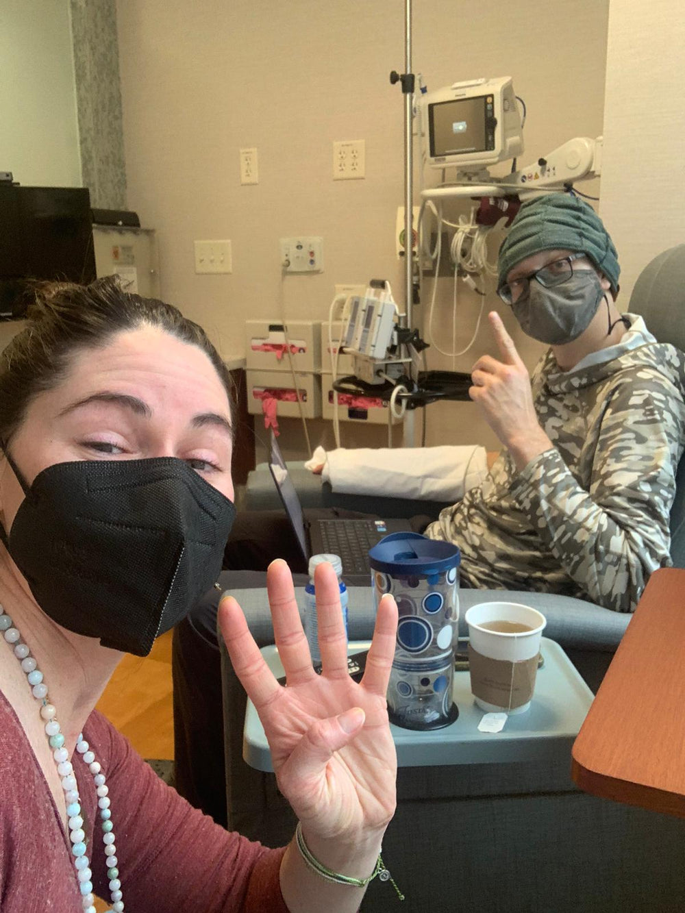 A lifestyle scene in a hospital or treatment room showing two people wearing face masks. In the foreground, a woman in a maroon top and light blue beaded necklace holds up five fingers.