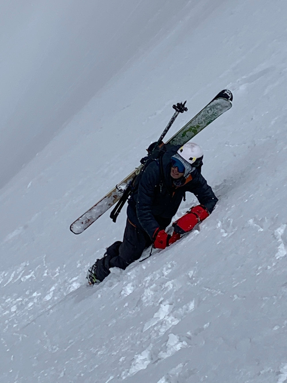 A skier in a dark jacket and pants, wearing a white helmet and goggles, struggles down a steep snowy slope while carrying skis and ski poles on their back. Bright red gloves are visible.