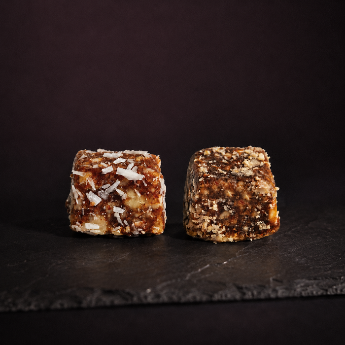 Two small square pieces of dense nut and coconut energy candy or protein bites standing upright on a dark slate surface against a dark brown background.